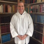 Smiling man in white postulant's smock standing in front of bookcase.