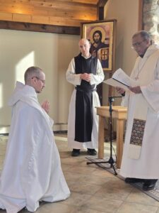 Brother Luke kneels in front of abbot Gerard in clothing ceremony at Genesee Abbey. Novice Director Br. Isaac stands in the background before an icon of Christ Pantocrator.