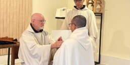 Brother Simon kneels before Abbot Paul-Mark as he makes his profession of vows. Another monk holds the vows book.