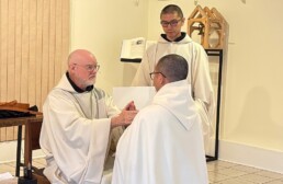 Brother Simon kneels before Abbot Paul-Mark as he makes his profession of vows. Another monk holds the vows book.